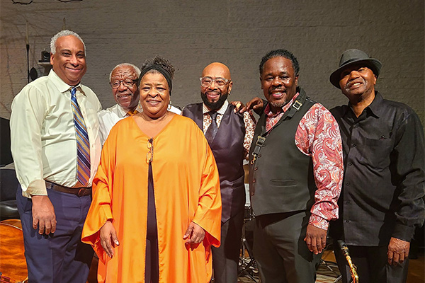Six smiling adults pose near musical instruments.