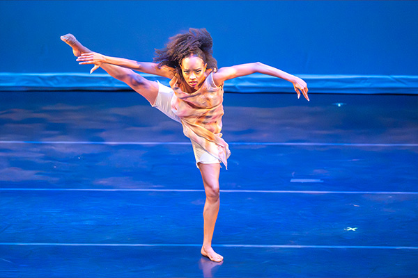 A woman dances barefoot on a stage, striking a dramatic pose with one leg high in the air.