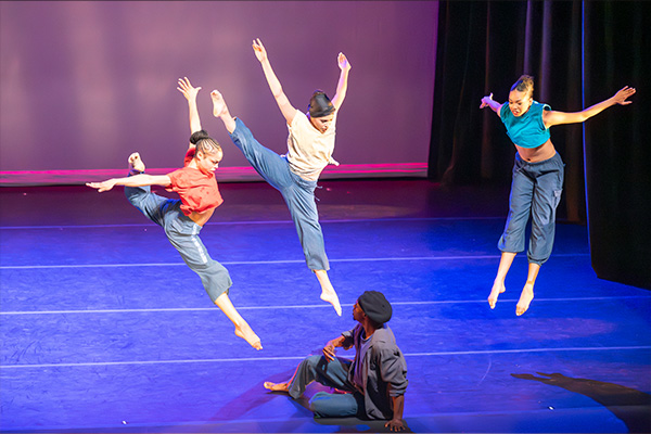 Four dancers perform on a stage. Three are leaping in midair, while one sits on the stage.