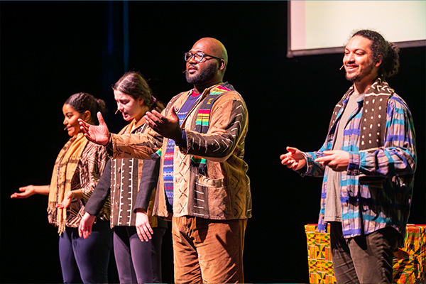 Four performers in patterned vests and scarves stand on a stage, addressing the audience.