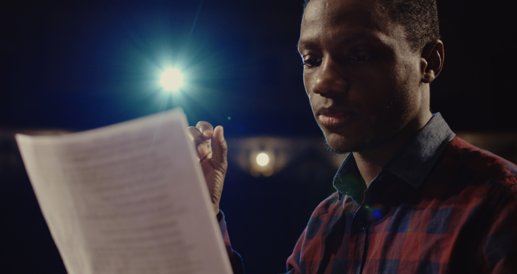 Young adult male in theatrical setting staring at a piece of paper with a stage light shining in the background.