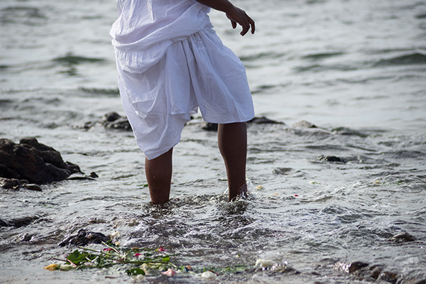 A person wearing white, wading into dark water. Flowers are washed up at the water's edge.