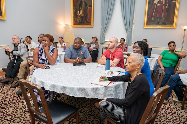 Audience members seated at round tables during a book talk in a historic room, listening to a speaker presentation.