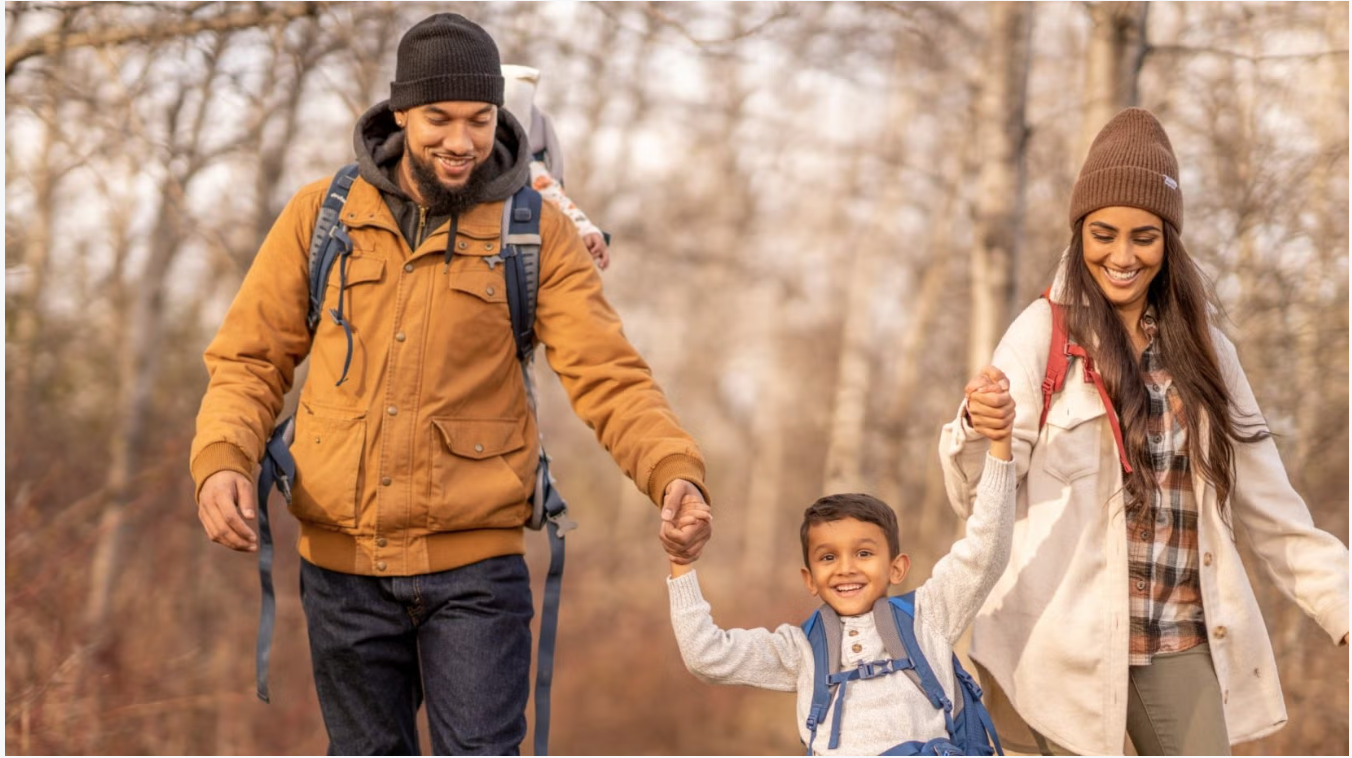 A smiling mother, father carrying a baby on his back, and young boy holding hands while walking through a wooded area.