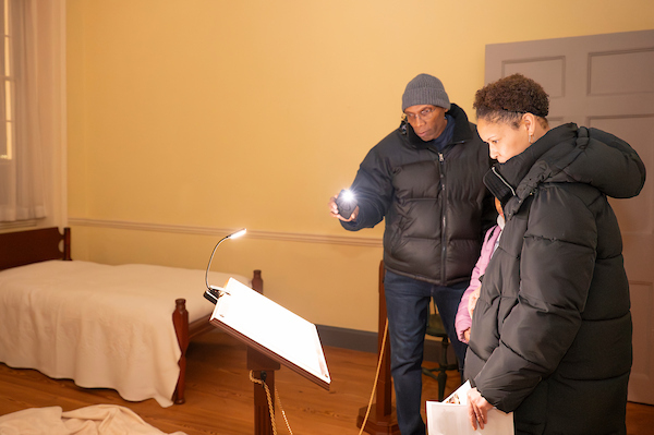 Visitors explore a historic bedroom exhibit by lamplight during a candlelight tour of a historic house.