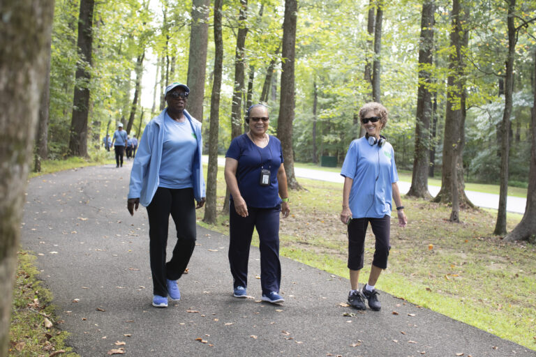 Three smiling older women wearing athletic clothing walking together on a paved path through a wooded park.