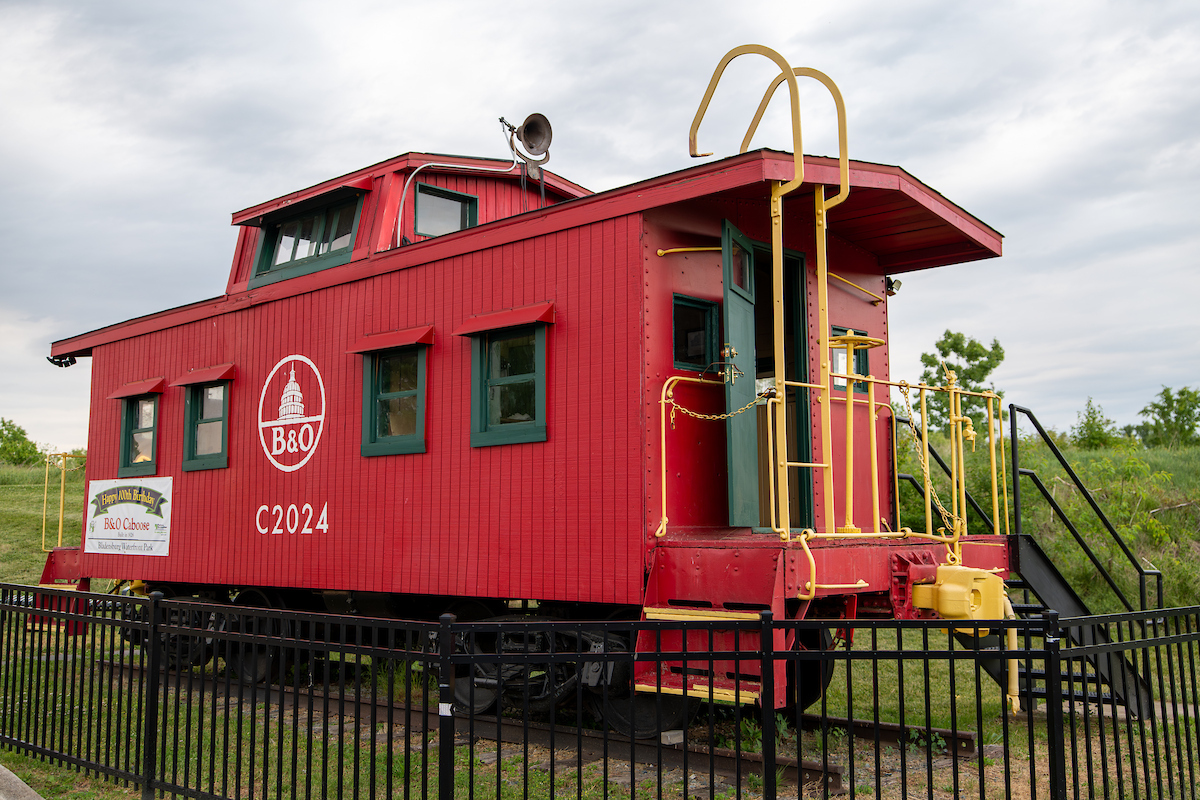 A red caboose sits on a short length of railroad track, with a metal stairway for access. A sign on the side reads B&O Caboose.