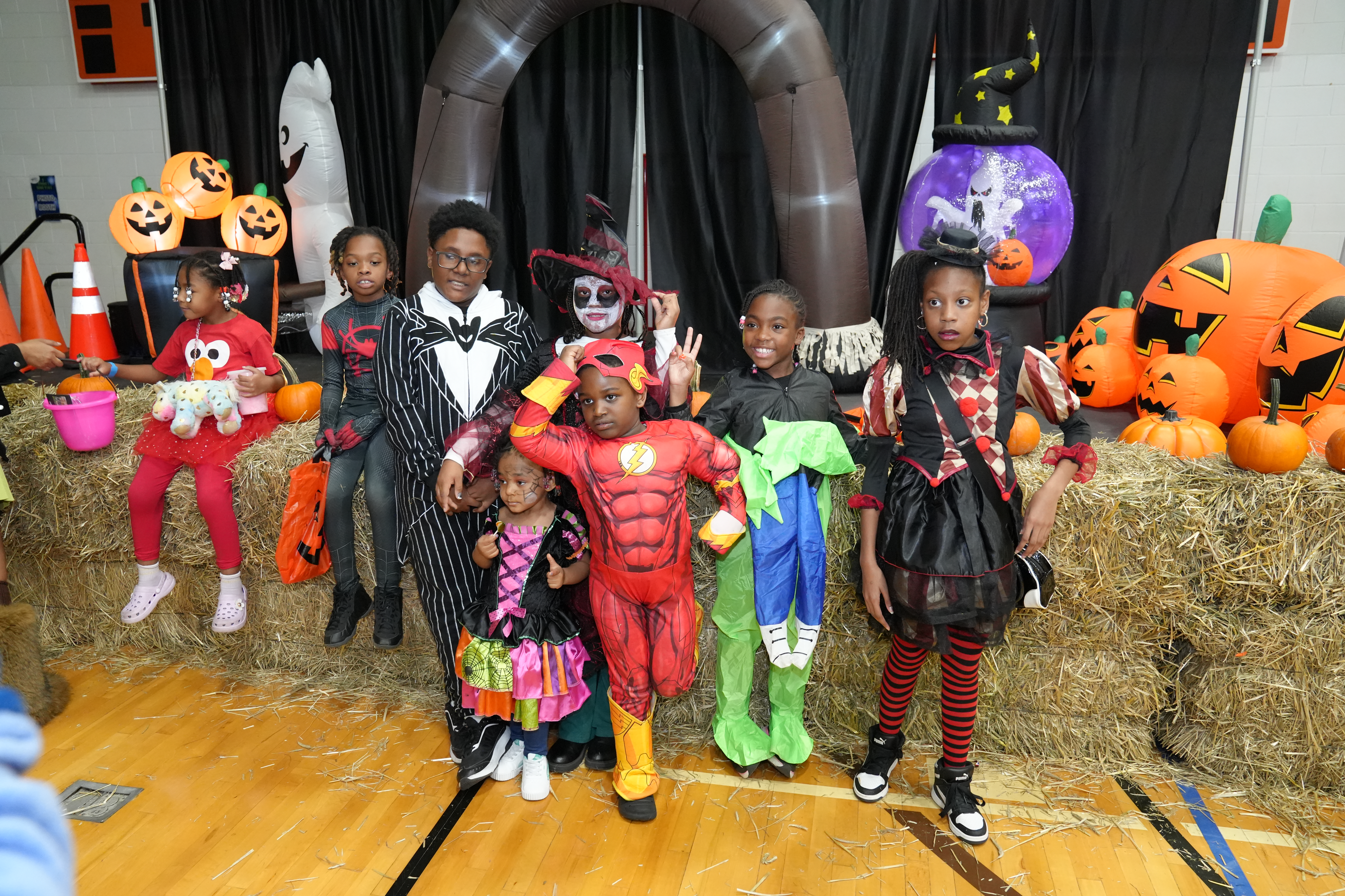 Children in Halloween costumes pose on hay bales beside inflatable pumpkins, a ghost, and festive decorations at an indoor Halloween celebration.