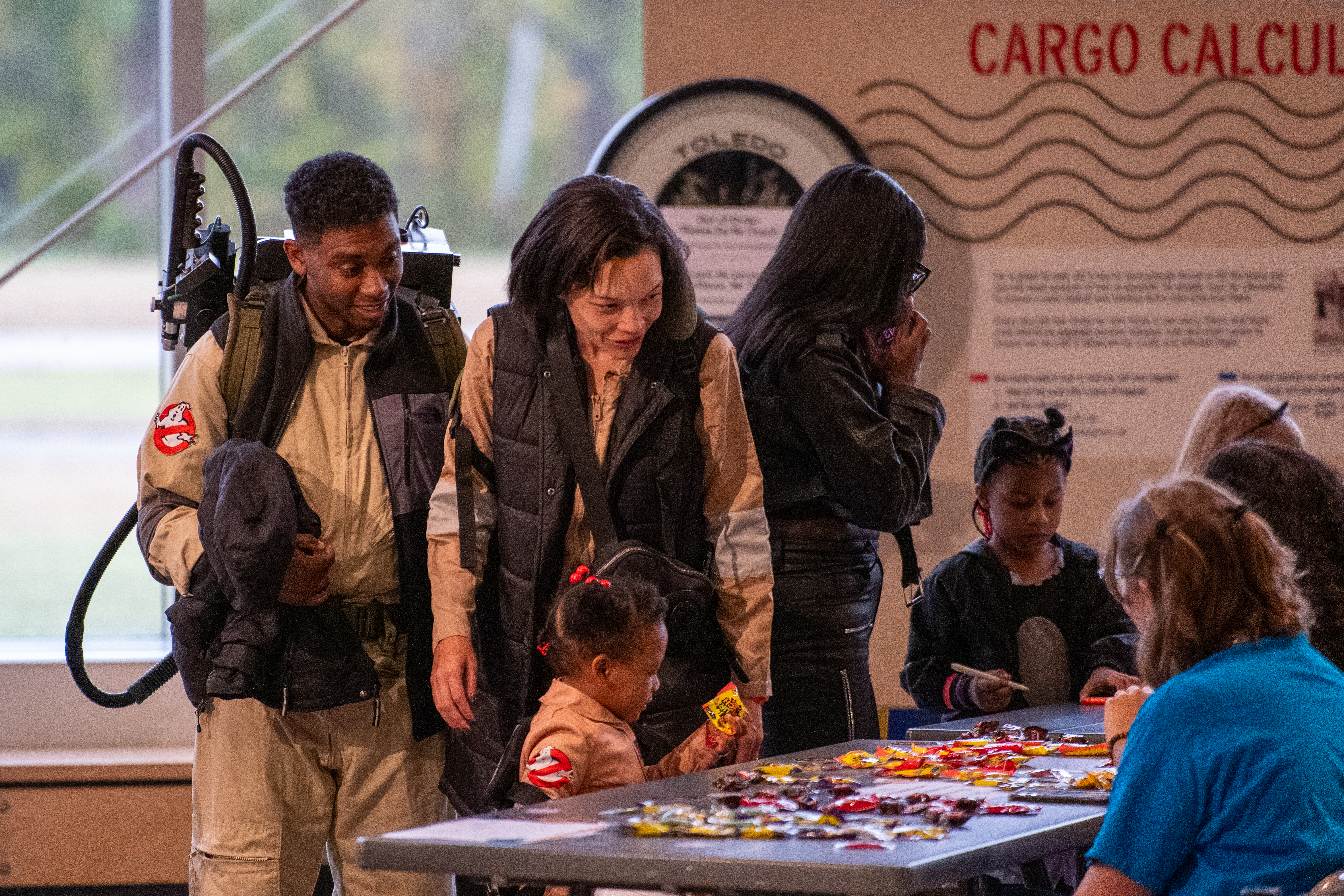 Visitors in costume gather around a candy table at a museum event, with one person wearing a Ghostbusters-style outfit and backpack prop.