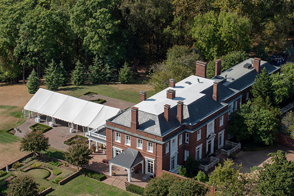 Aerial view of a large, historic, red brick manor house with a white event tent on the back patio and a garden on one side.