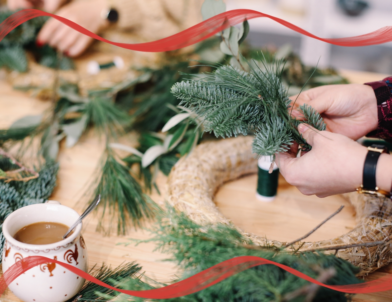 Close-up of hands crafting a holiday wreath with evergreen branches, straw base, and wire next to a hot cup of coffee.