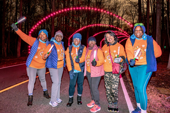 A group of women wearing matching orange shirts and winer gear pose in front of an outdoor light display at a park.